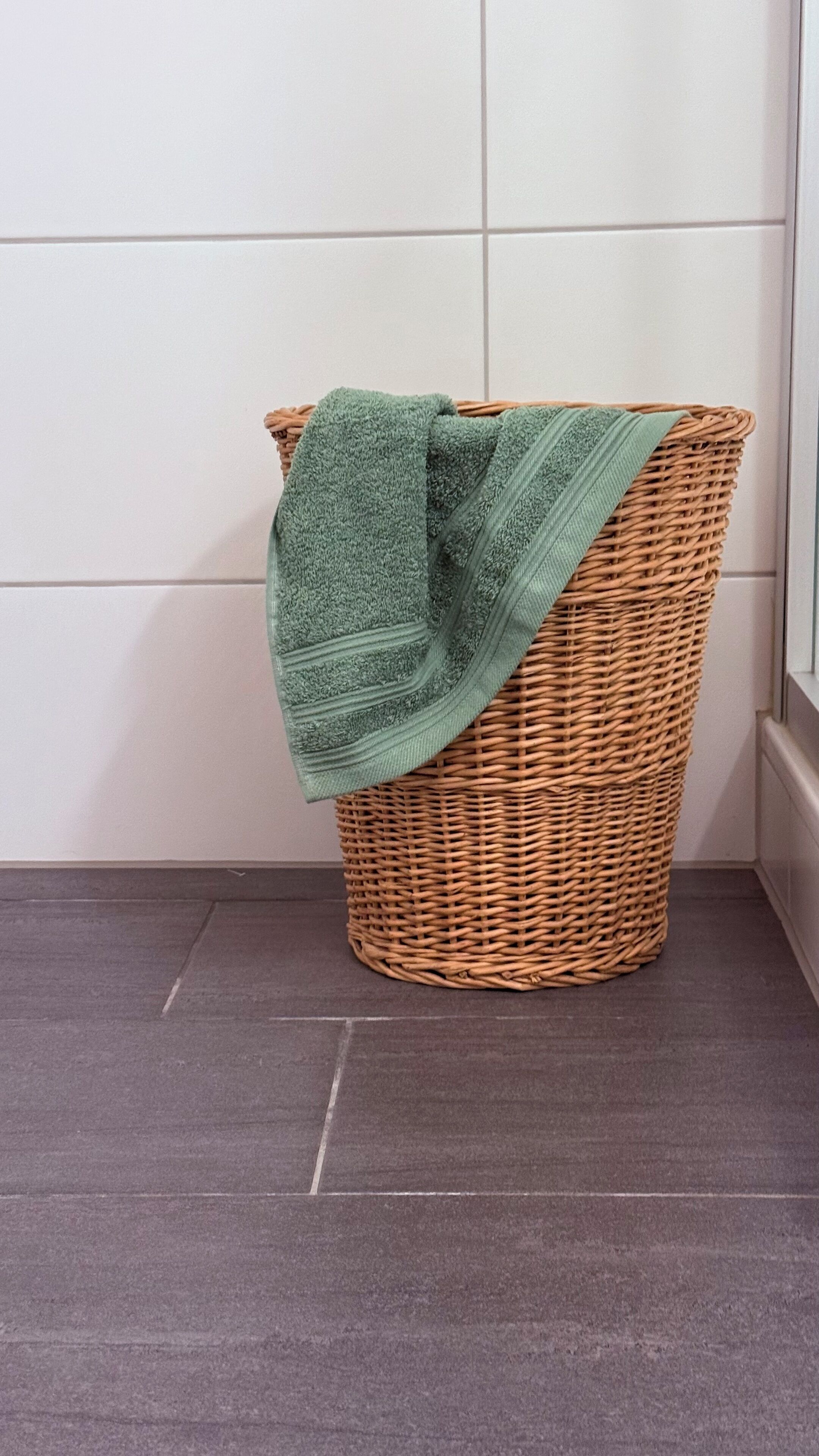 A wicker laundry basket with a green towel on a tiled floor.