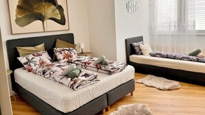 Een moderne slaapkamer met een groot bed, een kleinere bank, een decoratief wandobject en een raam met jaloezieën.