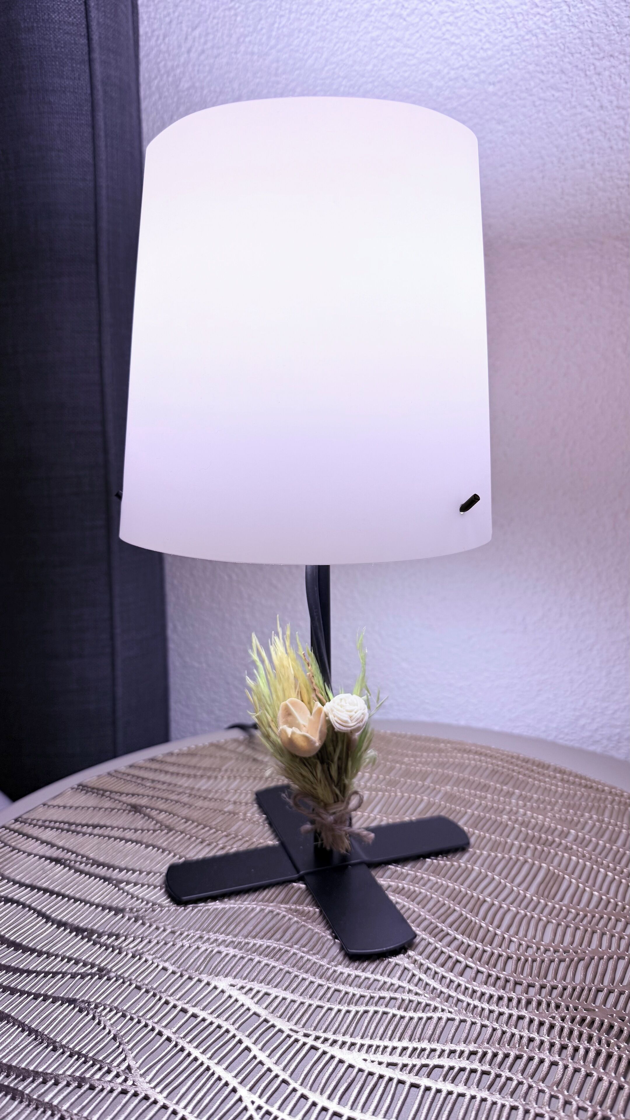 Een bureaulamp met een witte kap en een decoratieve bloemstelling op een geweven bijzettafeltje.