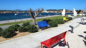 A coastal walkway with benches, a tree, and a clear view of the sea and distant buildings. - La Delfina by Interhome (Golfo Aranci)