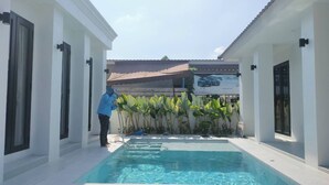 A swimming pool area with a person standing by the pool, surrounded by white buildings and a banner in the background.