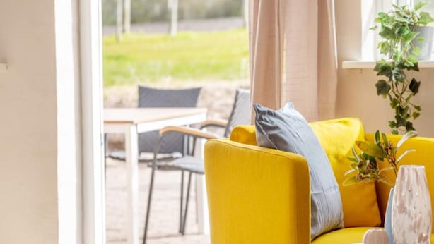 Un divano giallo brillante con un cuscino grigio, un vaso bianco con fiori e una pianta in vaso su un davanzale.