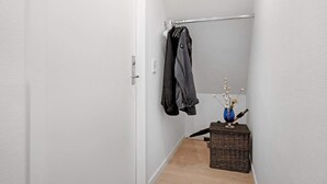 Un couloir étroit avec une porte blanche, un porte-manteau avec des vestes et un panier en osier contenant un vase de fleurs séchées.