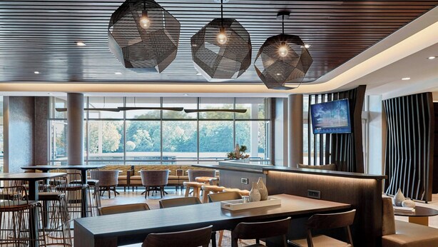Ein modernes Restaurant mit einem langen Gemeinschaftstisch, verschiedenen Sitzgelegenheiten und einem großen Fenster mit Blick ins Freie.