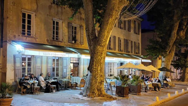 An outdoor café with tables and chairs under a tree, surrounded by buildings with signs for restaurants. - "Sur Place" 4-12 people. Ideally suited for two or a large family. (Carcès)