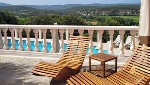 A balcony with wooden lounge chairs and a small table, overlooking a pool and a scenic view of hills and a village. - "Sur Place" 4-12 people. Ideally suited for two or a large family. (Carcès)
