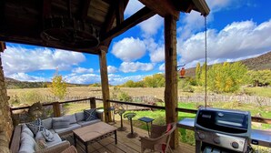 A covered outdoor area with a wooden deck, wicker furniture, a grill, and a view of a rural landscape. - Luxurious 2-bedroom cabin @ Canyon of the Ancients Guest Ranch in McElmo Canyon  (Cortez)