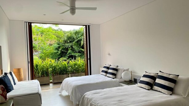 Ein modernes Schlafzimmer mit zwei Betten, einem Deckenventilator und einem großen Fenster mit Blick auf üppiges Grün.