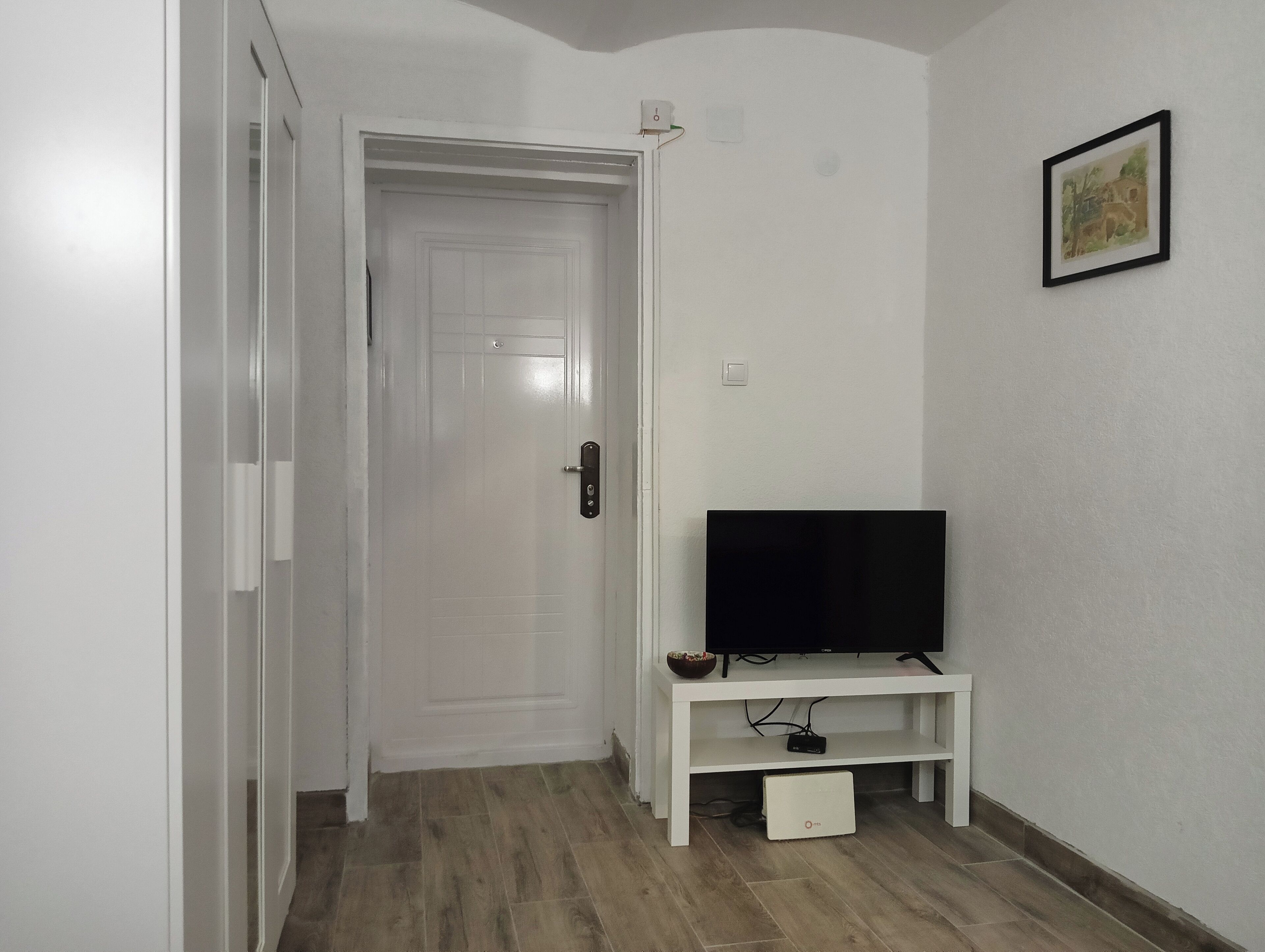 Een gang met een witte deur, een flatscreen televisie op een wit meubel en een lijst met een afbeelding aan de muur.