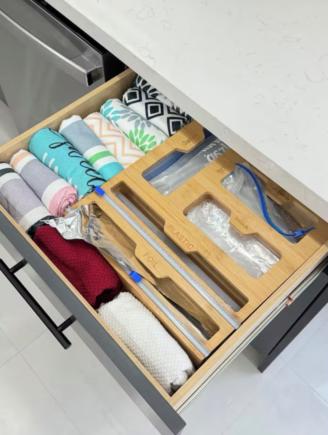 Eine Küchenschublade mit ordentlich gerollten Handtüchern, einer Plastiktüte und einer Holztrenneinlage mit Aufschriften 'FOIL' und 'PLASTIC'.