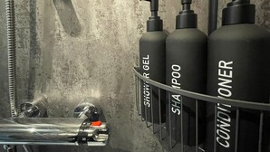Un baño con un estante metálico que contiene tres botellas etiquetadas como 'Shampoo Gel', 'Shampoo' y 'Conditioner'.