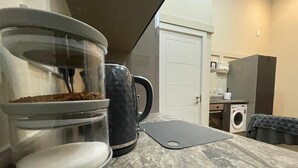 Una cocina con cafetera, hervidor y tabla de cortar sobre el mostrador.