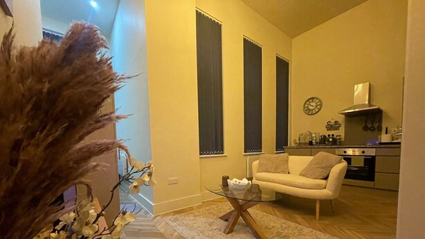 Una sala de estar con un sofá, una mesa de centro de vidrio y una planta decorativa. Se observa una cocina al fondo, con estufa y armarios.