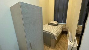 Un dormitorio compacto con armario, cama con colcha a cuadros, mesita de noche y ventana con cortinas.