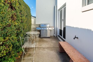 Eine Terrasse mit Grill, einem Metalltisch und Stühlen, einer Hecke und einer weißen Mauer.