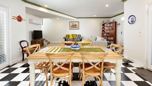A dining area with a checkered floor, a wooden table, and chairs. There is a bookshelf and a television in the background.