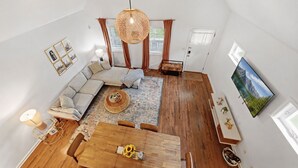 A spacious living area with a large sofa, a wooden coffee table, and a dining table. There is a hanging light fixture and a wall-mounted TV. - Coffee Lover's Paradise - Heart of Tremont (Cleveland)