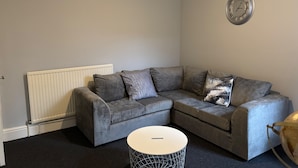A modern living room with a grey sectional sofa, a white radiator, a round coffee table, and a wall clock. - Oxford-house-sleeps 7-parking-garden (Oxford)