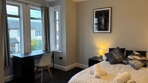 A hotel bedroom with a bed, a desk, and a chair. There is a window with curtains, a bedside lamp, and a framed picture on the wall. - Oxford-house-sleeps 7-parking-garden (Oxford)