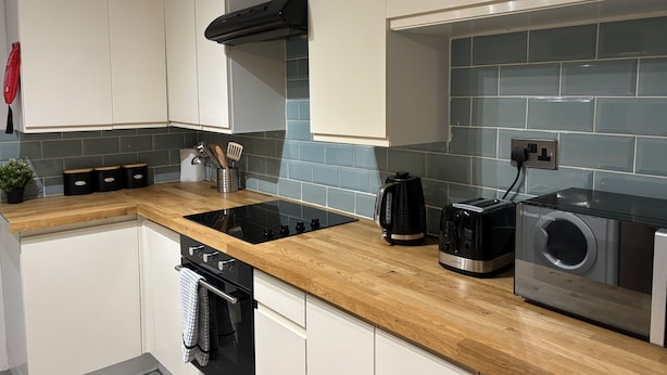 A modern kitchen with white cabinets, a wooden countertop, a black range hood, a kettle, a toaster, and a washing machine. - Oxford-house-sleeps 7-parking-garden (Oxford)