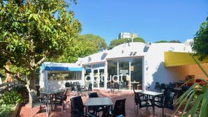 Zona de cafeteria a l'aire lliure amb taules i cadires, un edifici amb façana de vidre i un cel blau i clar.