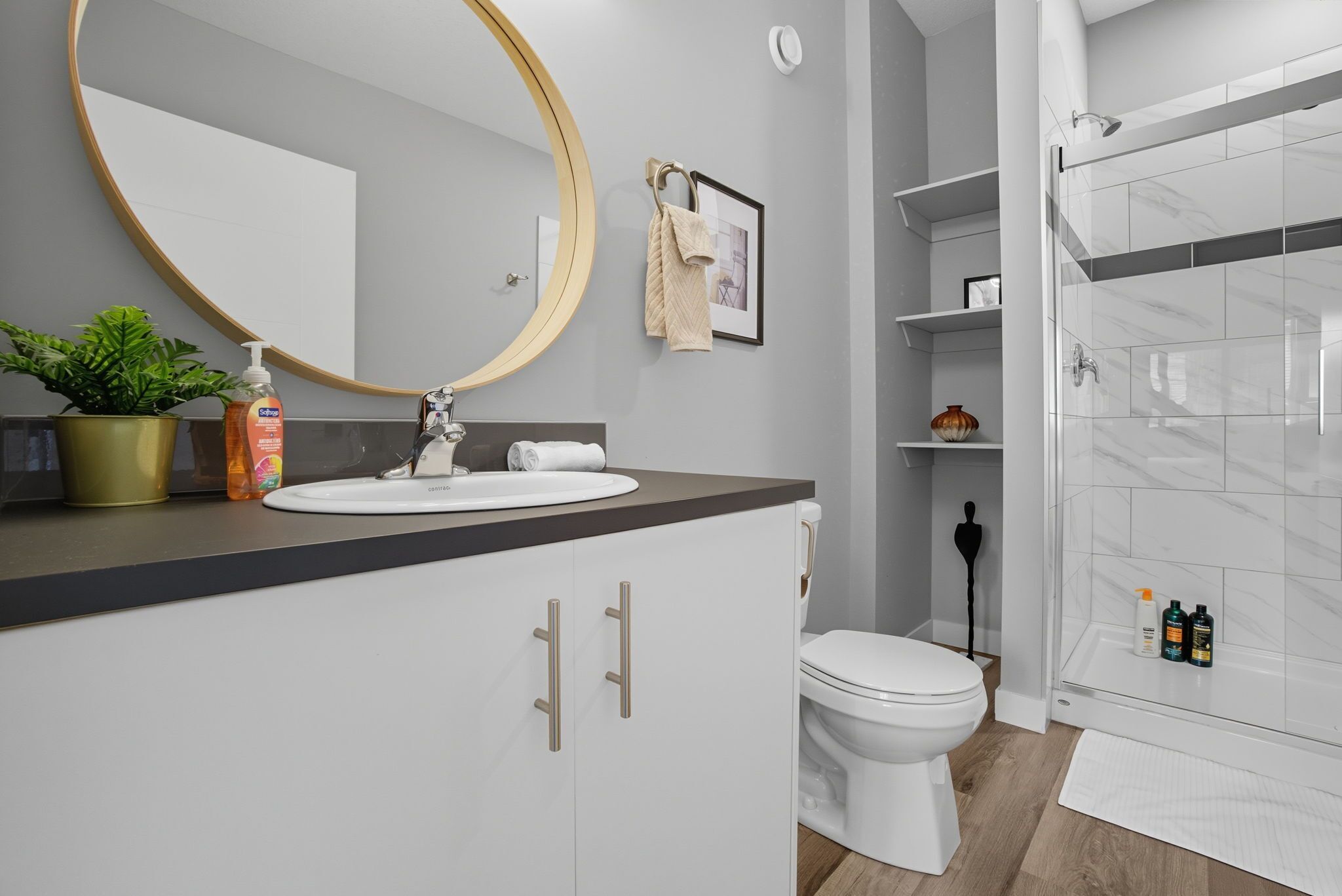 A modern bathroom with a white sink, mirror, and toilet. There is a shower area with a glass door and a potted plant on the counter.