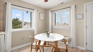 Una habitación con una mesa redonda, dos ventanas con persianas, un ventilador de techo y una puerta.