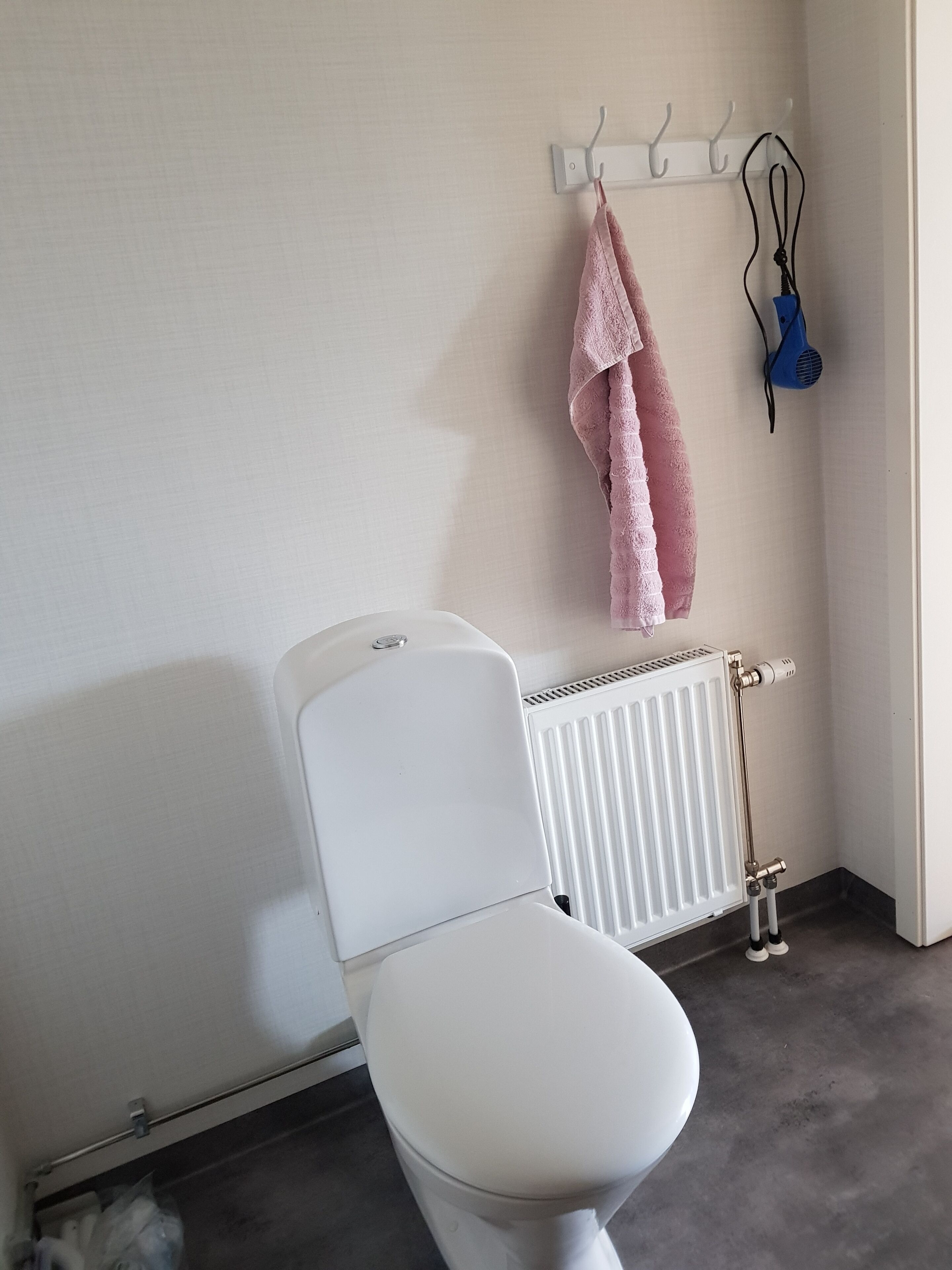 A white toilet with a radiator below it, a towel hanging on a hook, and a blue hairdryer on the wall.