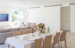 A modern dining area with a long table set for six, surrounded by beige chairs. A sofa and a dining table are positioned near a large window with a view of greenery.