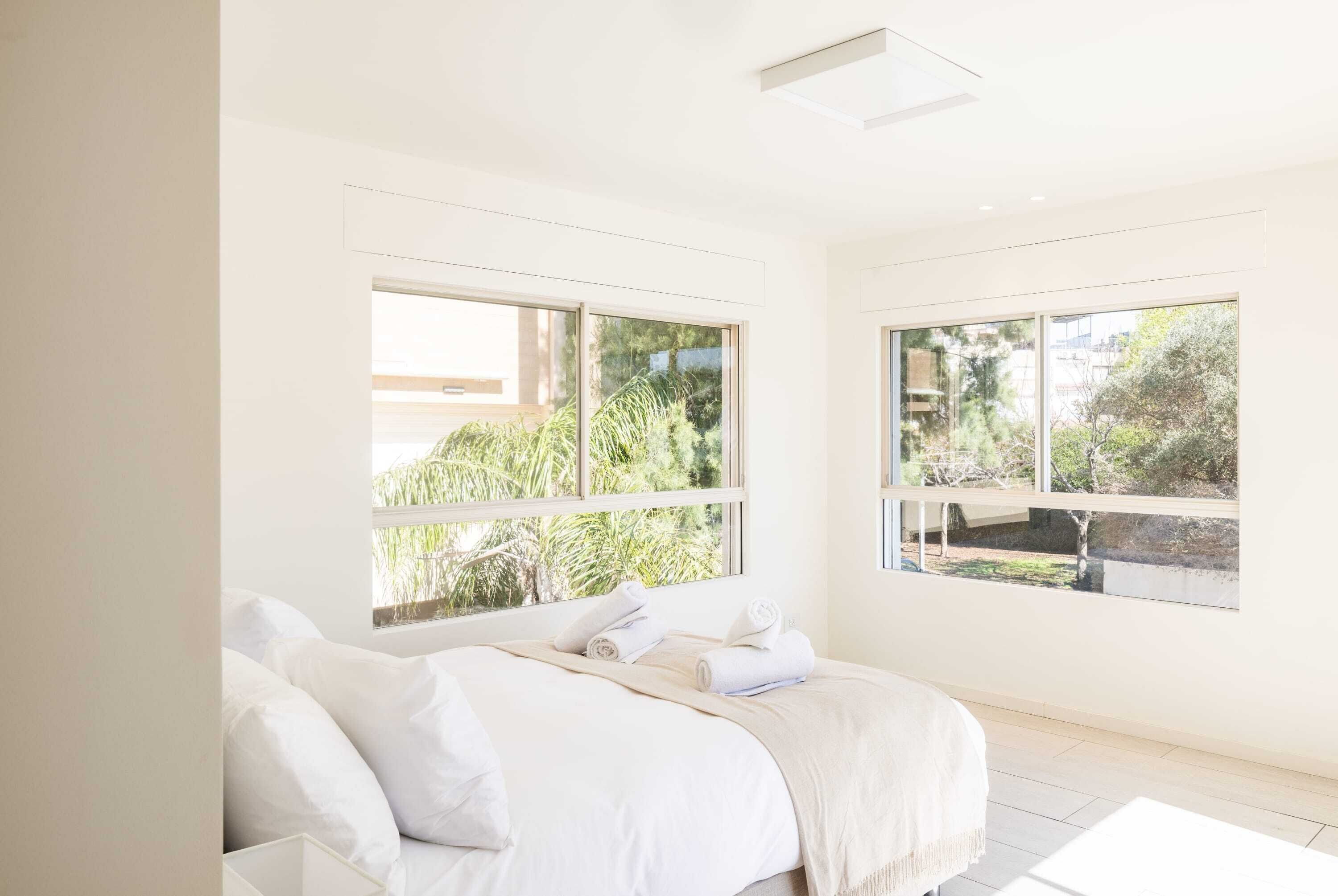 Una camera da letto luminosa e minimalista con un letto grande, due finestre e vista sul verde.