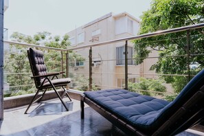 A balcony with a cushioned seating area and a chair, overlooking a residential building and greenery. - Carmelo  Gordon Sunset with MAMAD (Tel Aviv-Yafo)