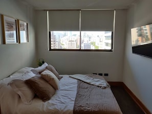 Un dormitorio con cama, ventana con persianas y vista a la ciudad.