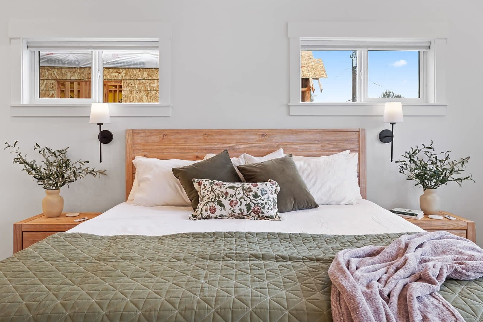 A bedroom with a wooden headboard, two bedside tables with lamps, a green quilted bedspread, and a view of a construction site through the window.