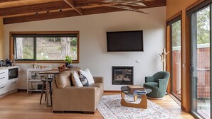 A modern living room with a wooden ceiling, a large window, a flat-screen TV, a fireplace, and a sliding glass door leading to an outdoor area. - Wedge-tailed Eagle Studio (Kettering)