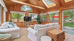 Une véranda spacieuse avec des murs et un plafond en bois, de grandes fenêtres, un ventilateur de plafond et un téléviseur.
