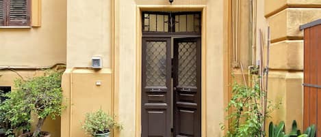 A yellow building with a dark brown door, potted plants, and a small lamp above the door.