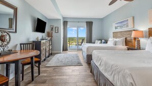 A hotel room with two beds, a desk, a television, and a balcony with a view.