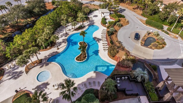 An aerial view of a resort with a large swimming pool, palm trees, and a winding road.