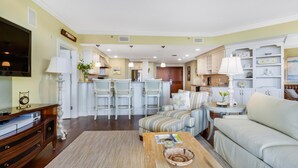 A living room with a sofa, armchairs, a coffee table, and a TV. There is a kitchen area with bar stools and a dining table.