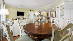 A dining area with a round wooden table, white chairs, and a television in the background.