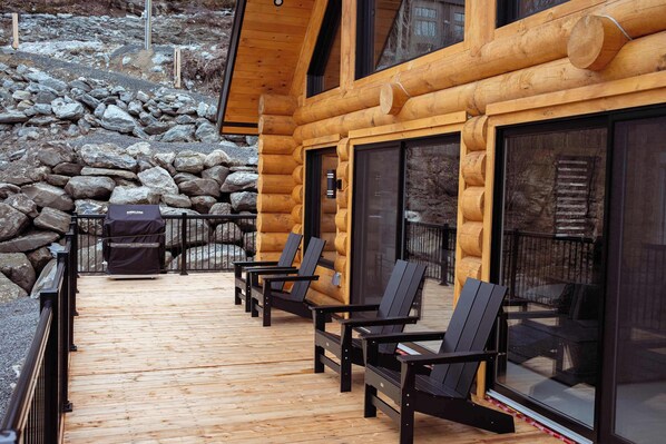 Una terraza de madera con sillas y una parrilla, un muro de piedra y una cabaña de troncos con ventanas amplias.
