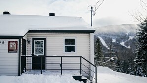 Ein schneebedecktes Gebäude mit einer Treppe zum Eingang, einem kleinen Fenster und Blick auf eine verschneite Berglandschaft.