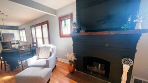 Ein Wohnzimmer mit Kamin, Holzböden, einer Essecke mit Tisch und Stühlen sowie einer Küche im Hintergrund.