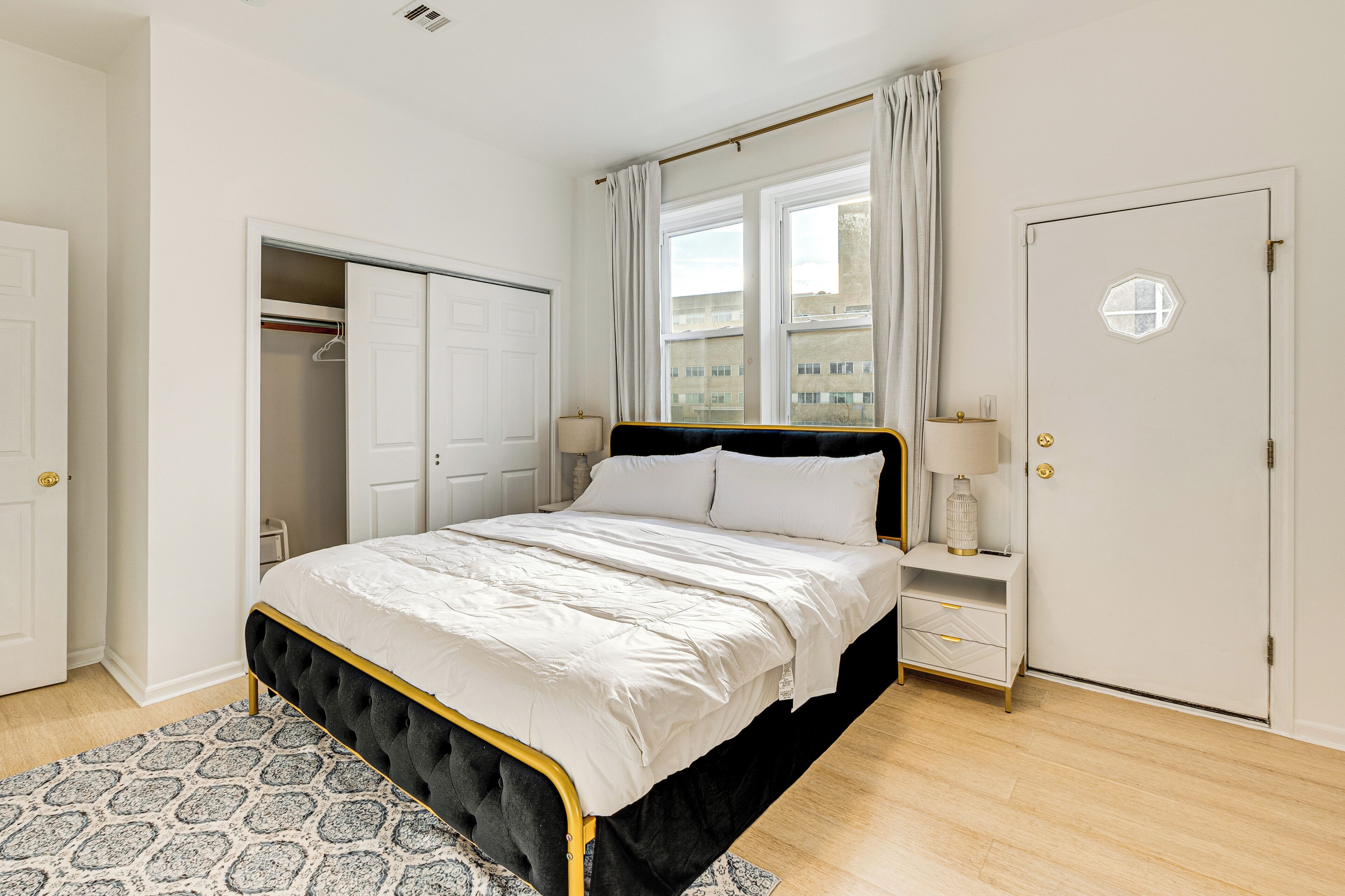 Un dormitorio con cama, armario, mesita de noche, lámpara y ventana con cortinas.