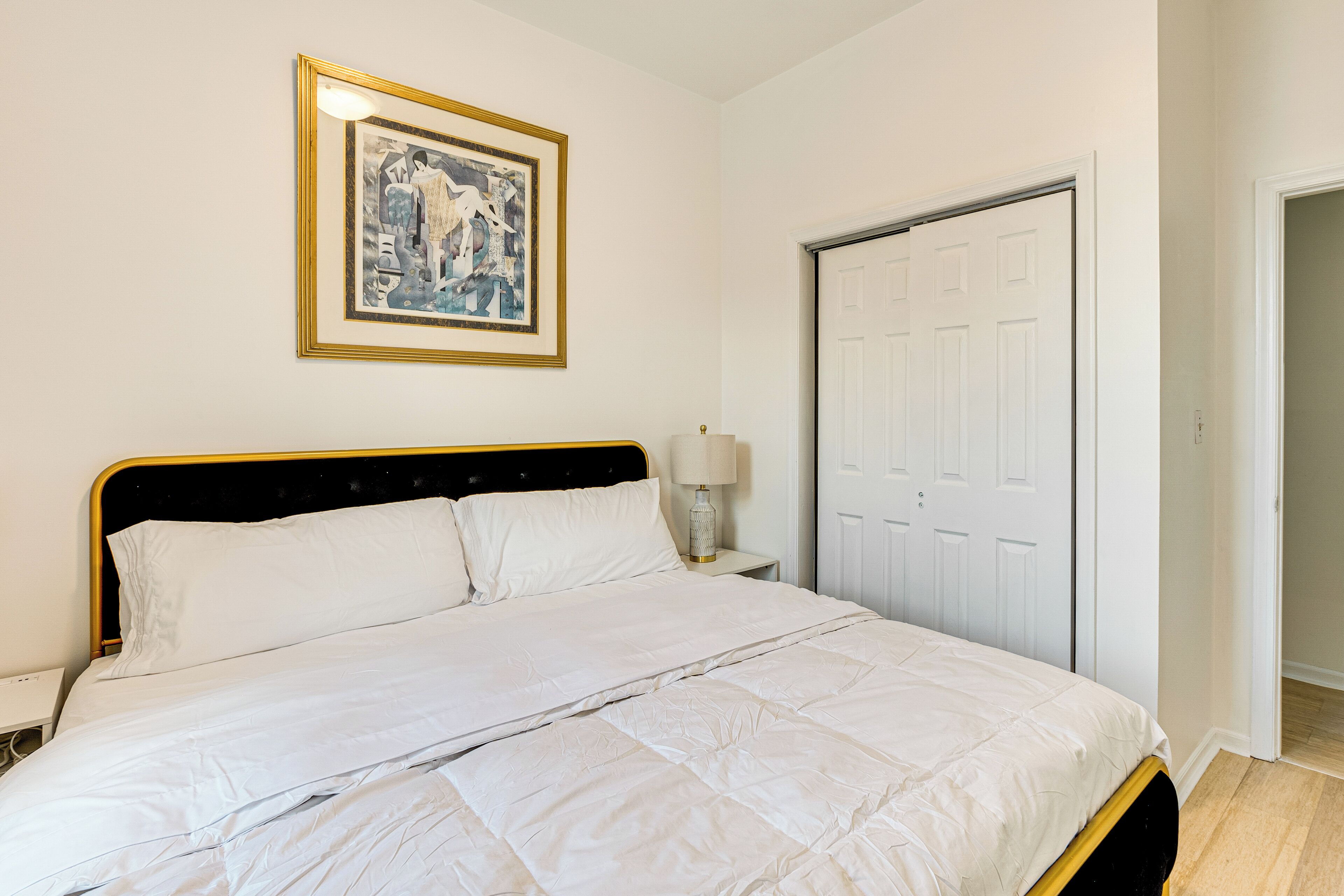Una cama bien hecha con ropa blanca, una obra de arte enmarcada encima y una puerta cerrada a la derecha.