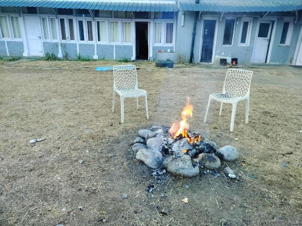 A fire pit with two white chairs in front, surrounded by a dry grassy area, with a building in the background.