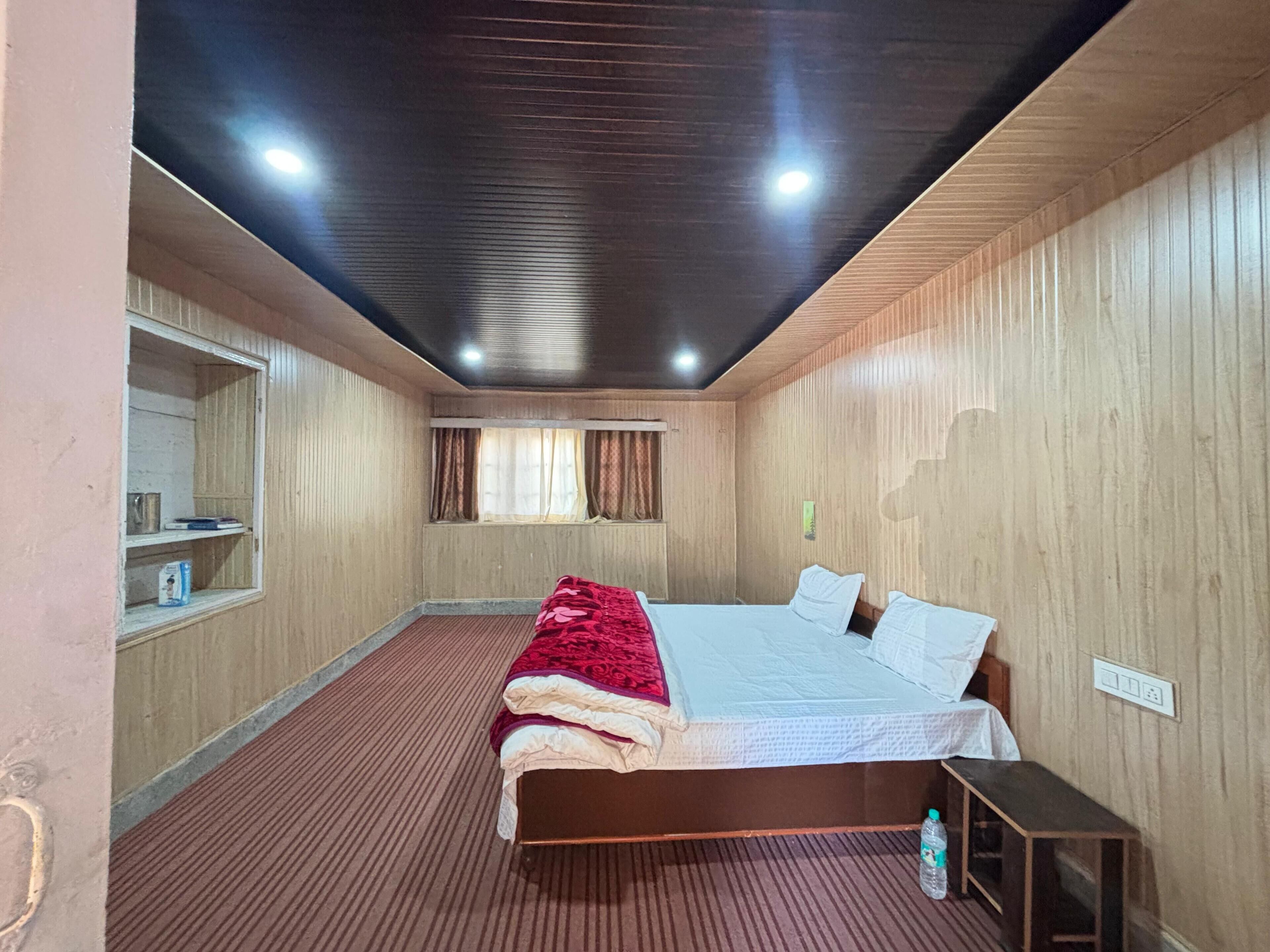 A hotel room with a wooden ceiling, a bed with a red blanket, a bedside table, and a window with curtains.