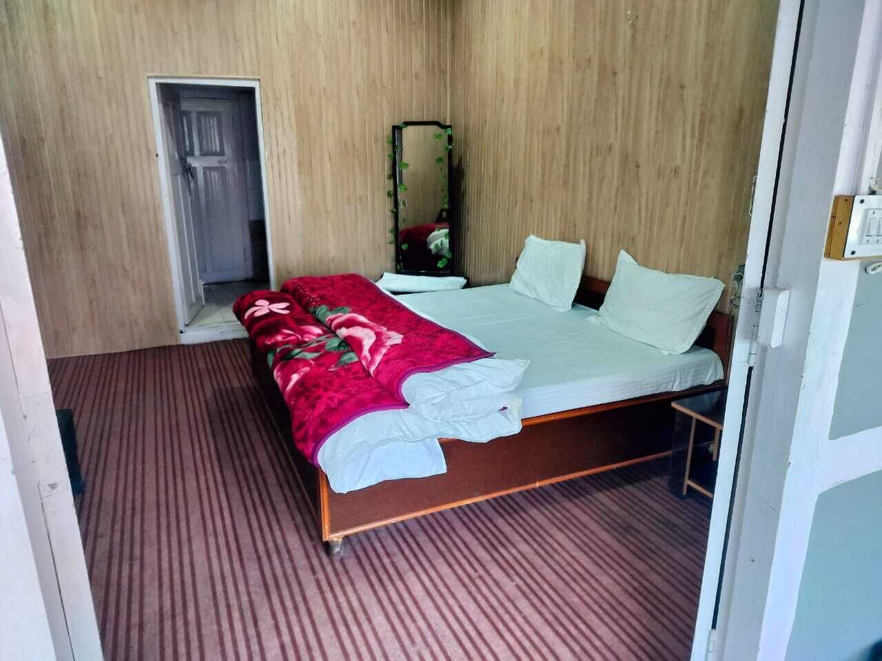 A single bed with a red blanket and white pillows, a wooden door, and a mirror on the wall.