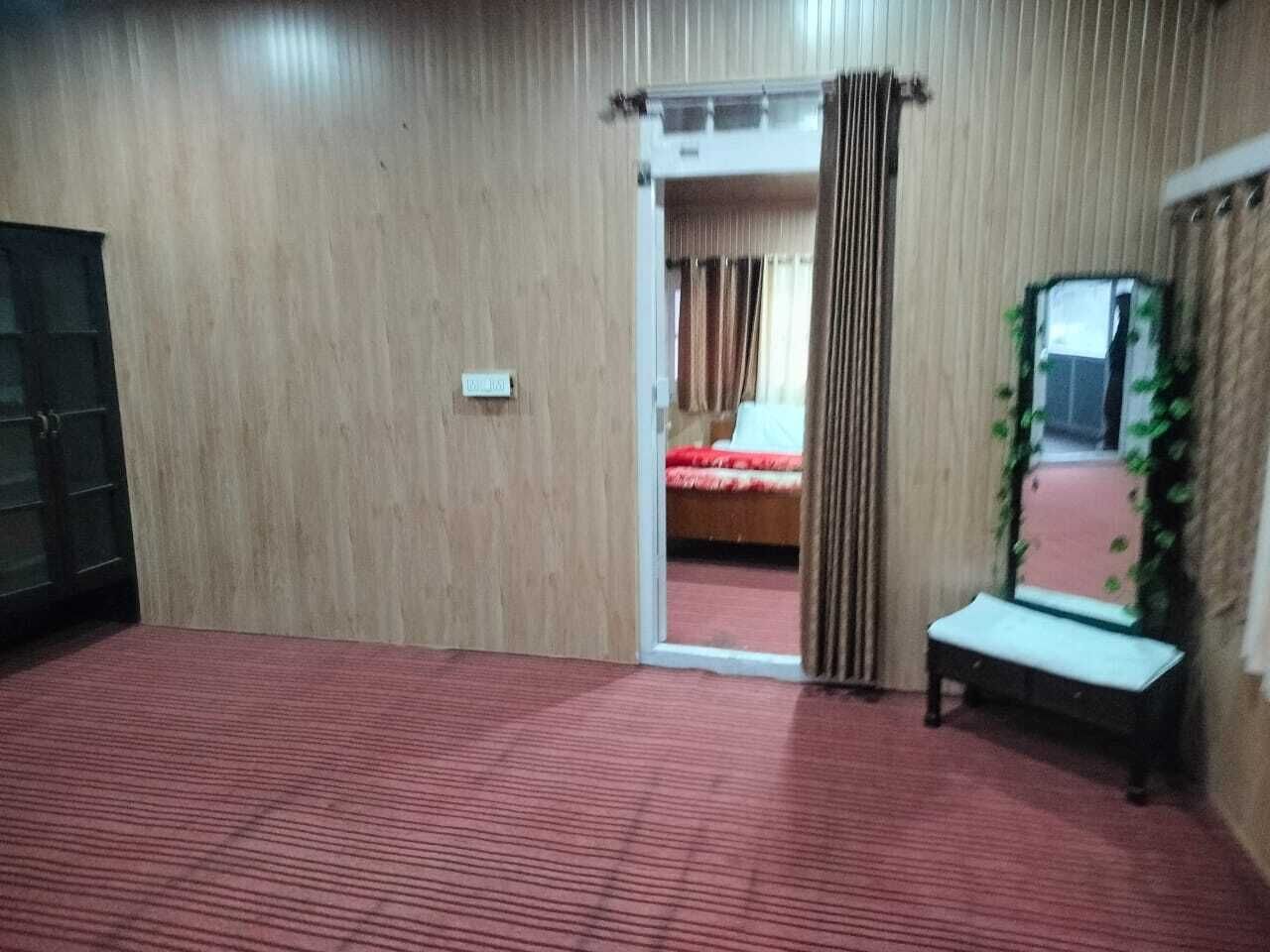 Habitació d'hotel amb una catifa vermella, una banca, un mirall i una porta que dona a un dormitori amb un llit i cortines.