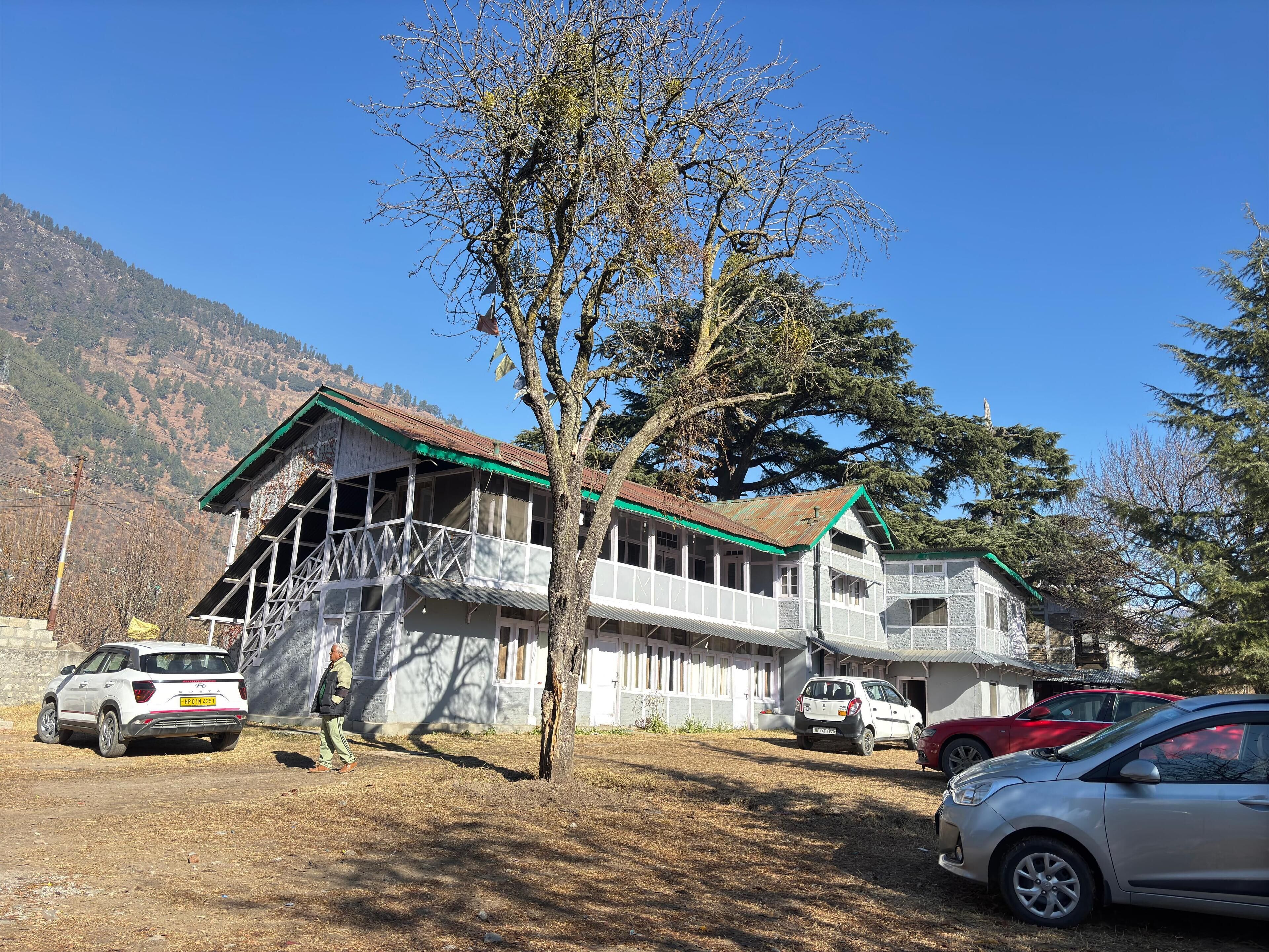 Un bâtiment à deux étages, doté d’un toit végétalisé et de murs blancs, entouré de voitures garées et d’un arbre.